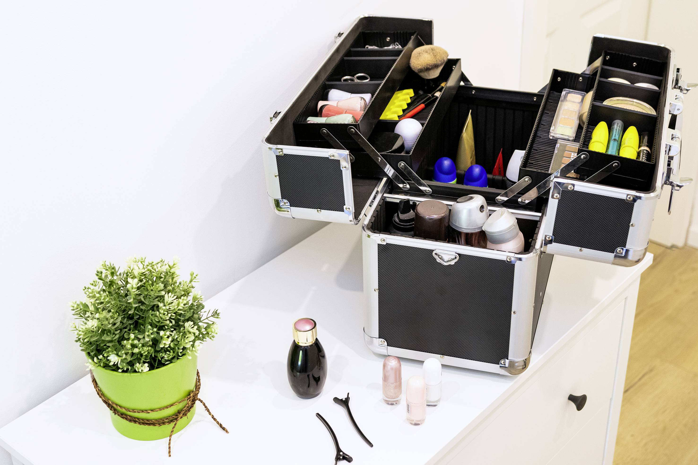 Nyitott sminktáska fehér asztalon, benne különböző kozmetikumokkal és eszközökkel. Mellette egy zöld növényt tartalmazó cserép áll.
