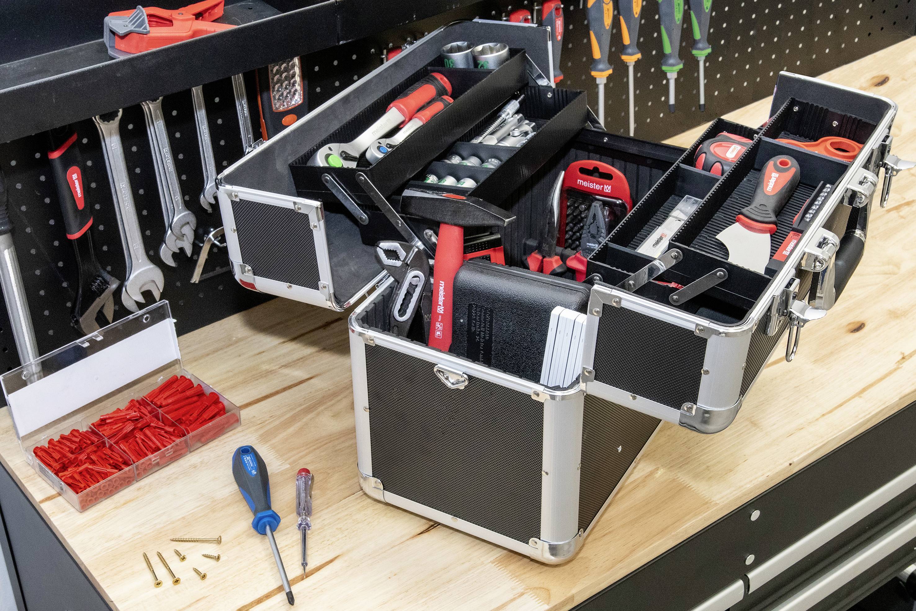 Egy nyitott szerszámosláda áll egy munkapadon. Különböző szerszámok, mint csavarhúzók és fogók láthatók. A háttérben további szerszámok lógnak a falon.