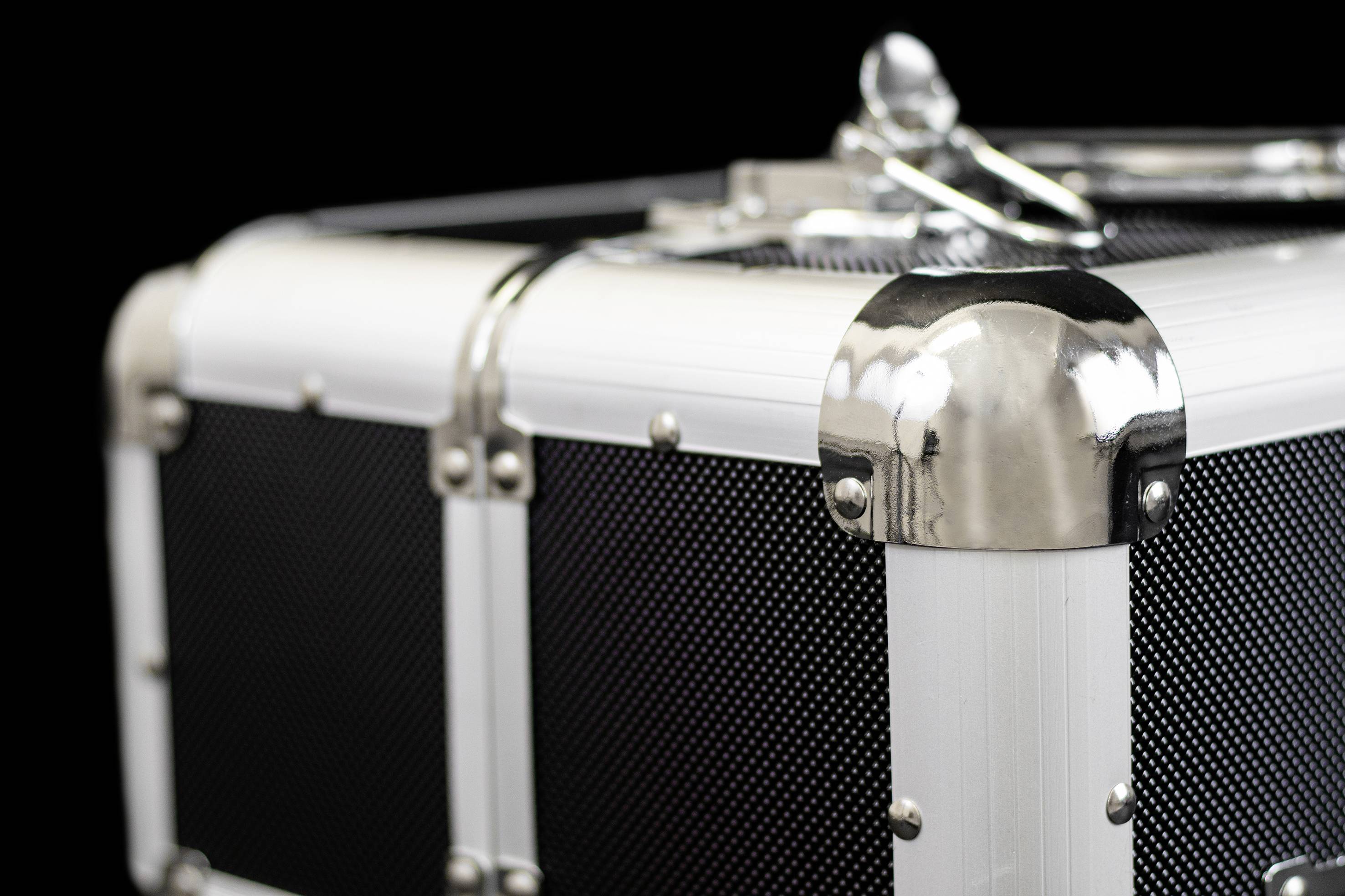 Egy ezüstös, megerősített fémtok fekete fogantyúval és fényes sarkokkal, fekete háttér előtt.