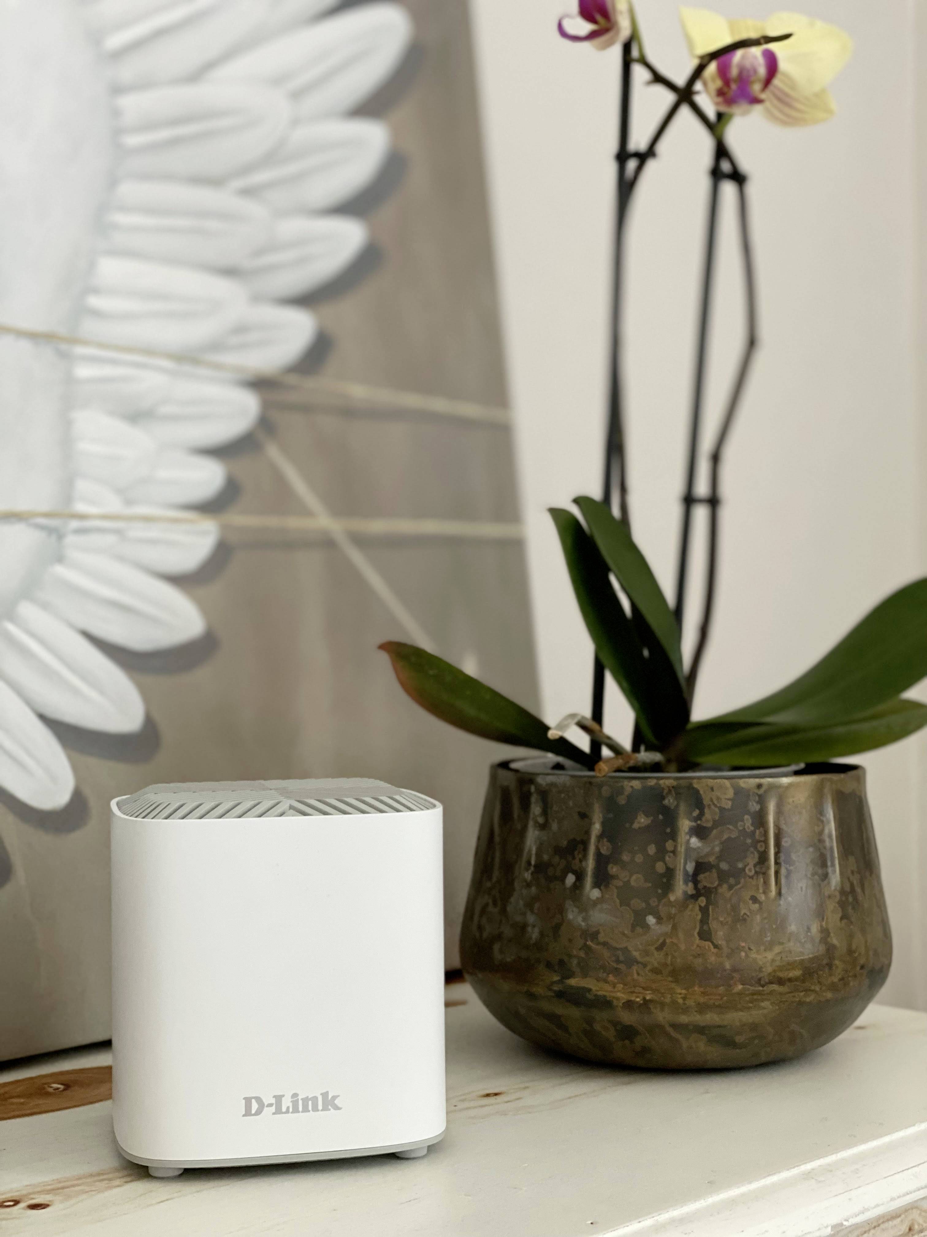 Egy fehér D-Link WLAN-router áll egy polcon egy barna cserépben lévő orchidea mellett. A háttérben egy absztrakt kép látható.