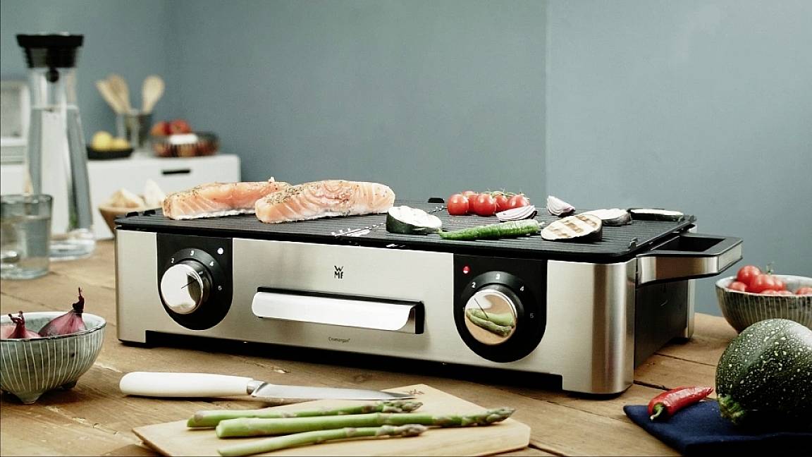 Egy elektromos asztali grill lazaccal, paradicsommal és spárgával a sütőfelületen fa asztalon áll. Konyhai eszközök láthatók a háttérben.
