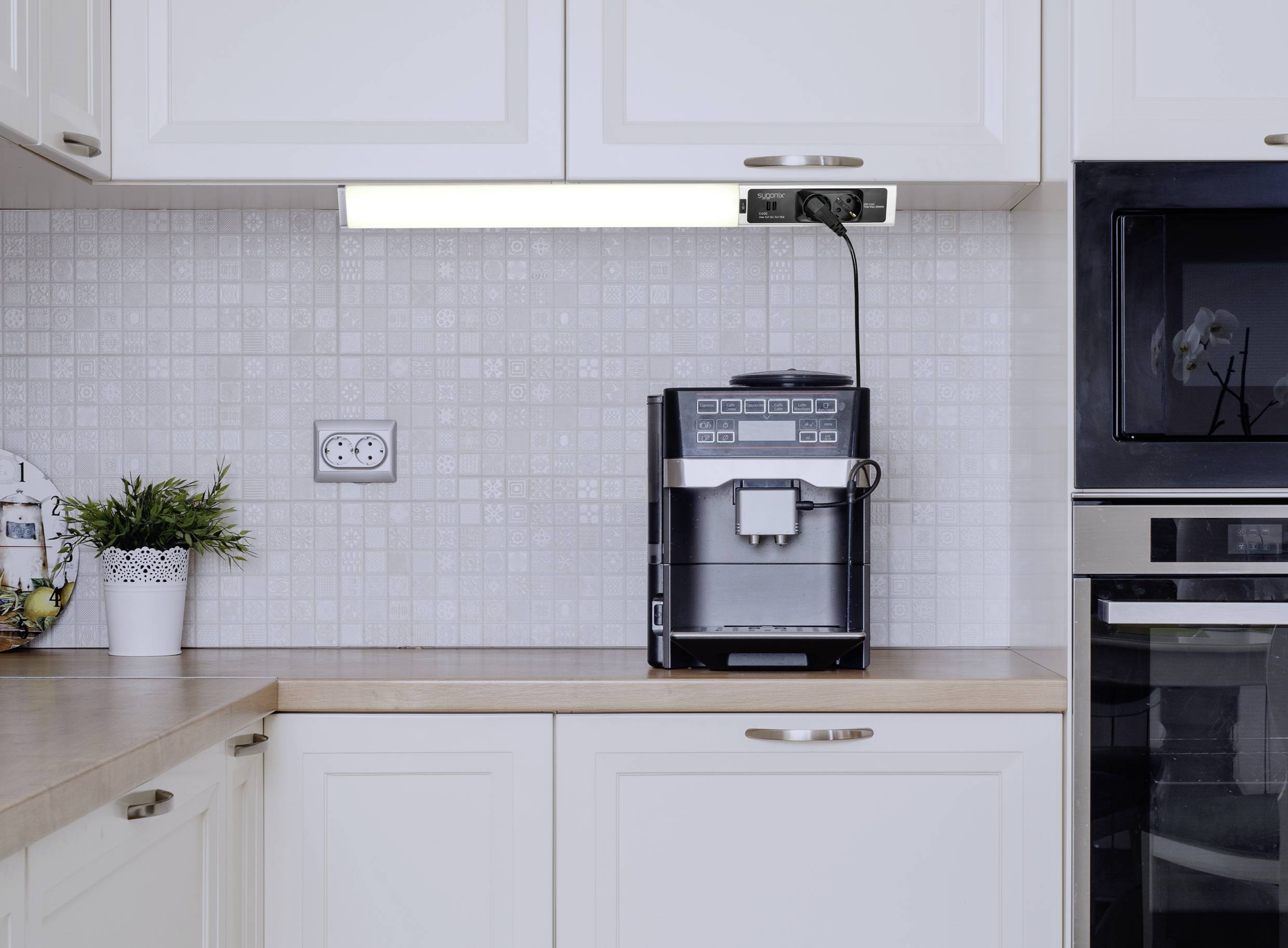Modern konyha fehér berendezéssel. Egy kávéfőző áll a munkafelületen egy felső szekrény alatt, mellette növények.