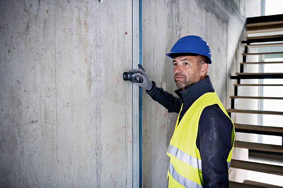 Egy munkás biztonsági mellényben és védősisakban egy eszközzel méri egy betonfalat egy épülő szerkezetben. A háttérben egy lépcső látható.