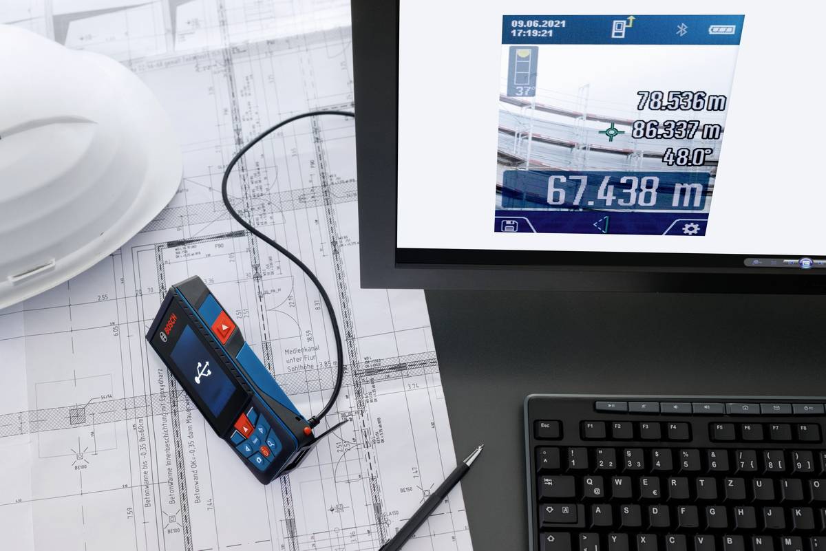 Egy digitális mérőkészülék '67,438 m' értéket mutat egy képernyőn, amelynek környezetében építési tervrajzok és egy védősisak láthatók. Egy monitor további mérési eredményeket jelenít meg.