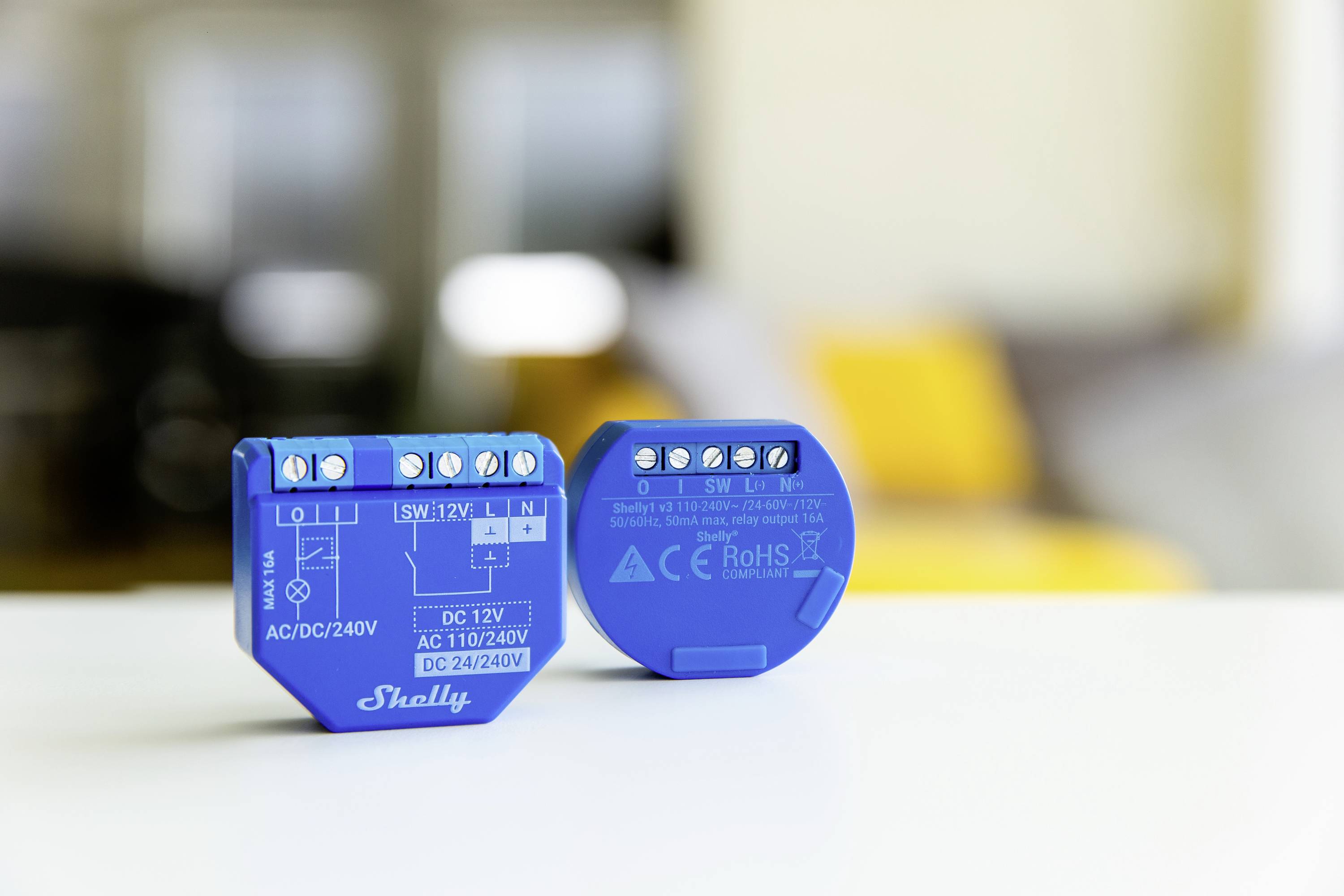 Két kék elektronikai modul egy asztalon, az egyik "Shelly" felirattal. A háttér elmosódott sárga és szürke árnyalatokkal.