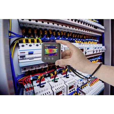 Egy kéz egy mérőműszert tart egy nyitott elosztószekrény előtt, amelynek kábelezése látható, elektromos mérések elvégzése céljából.