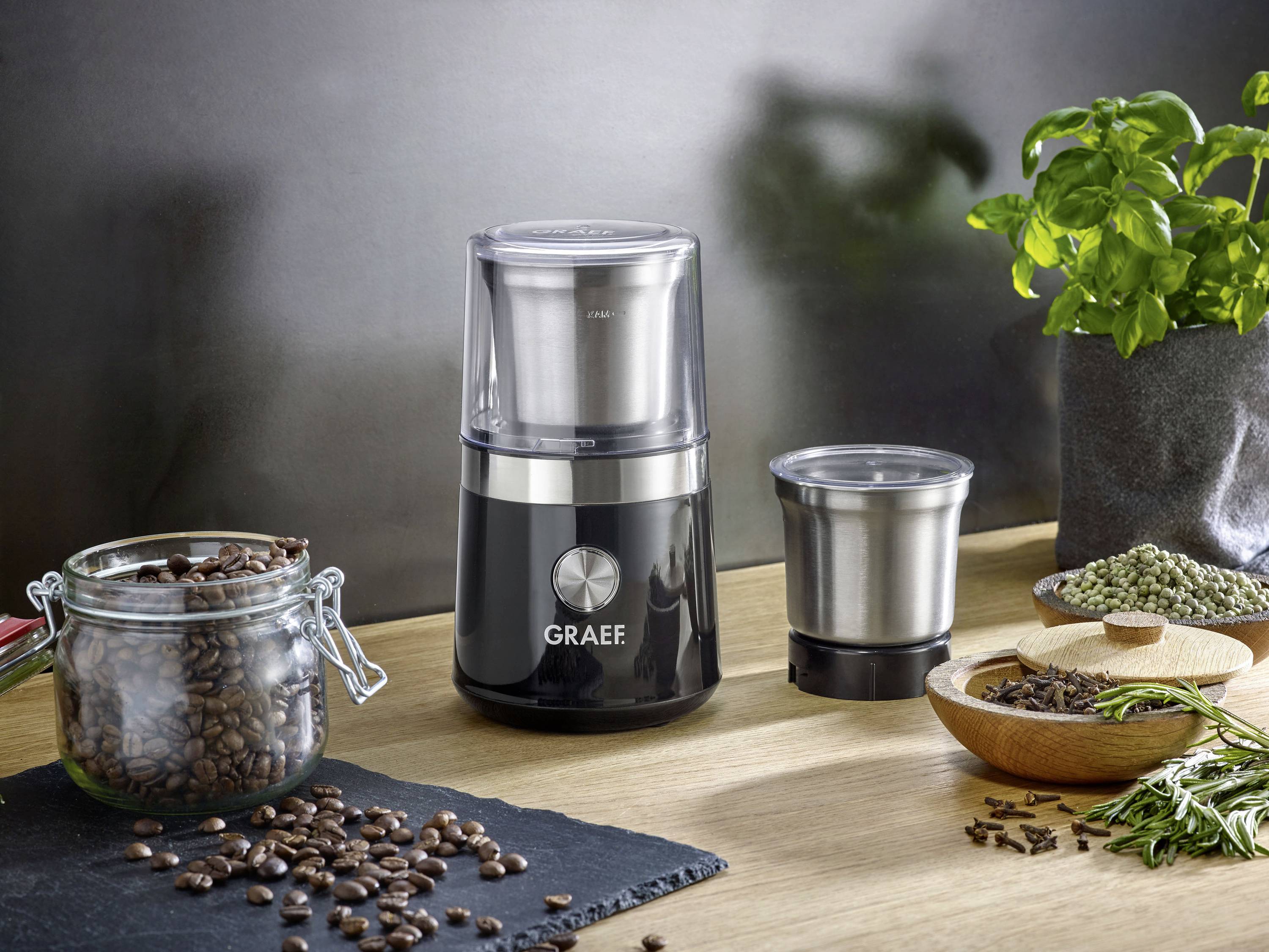 Elektromos kávédaráló egy asztalon, kávébabokkal, fűszerekkel és egy növénnyel mellette; modern és praktikus kialakítású.