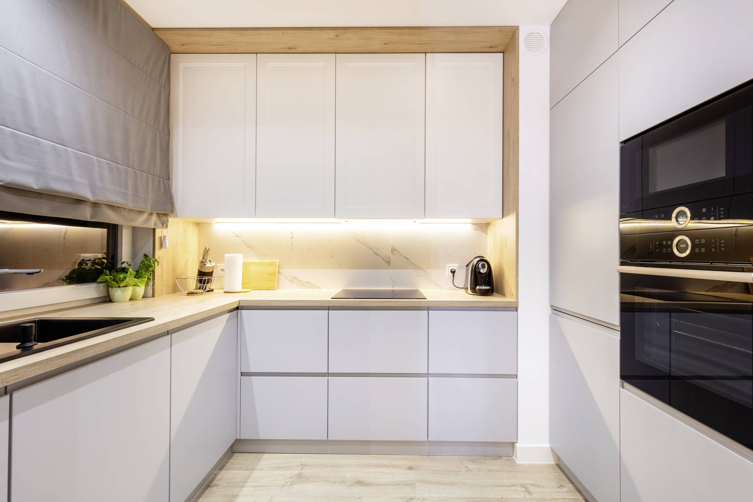 Egy modern konyha világos szekrényekkel, üvegkerámia főzőlappal, beépített sütővel, szürke munkafelületekkel és egy nagy ablakkal.