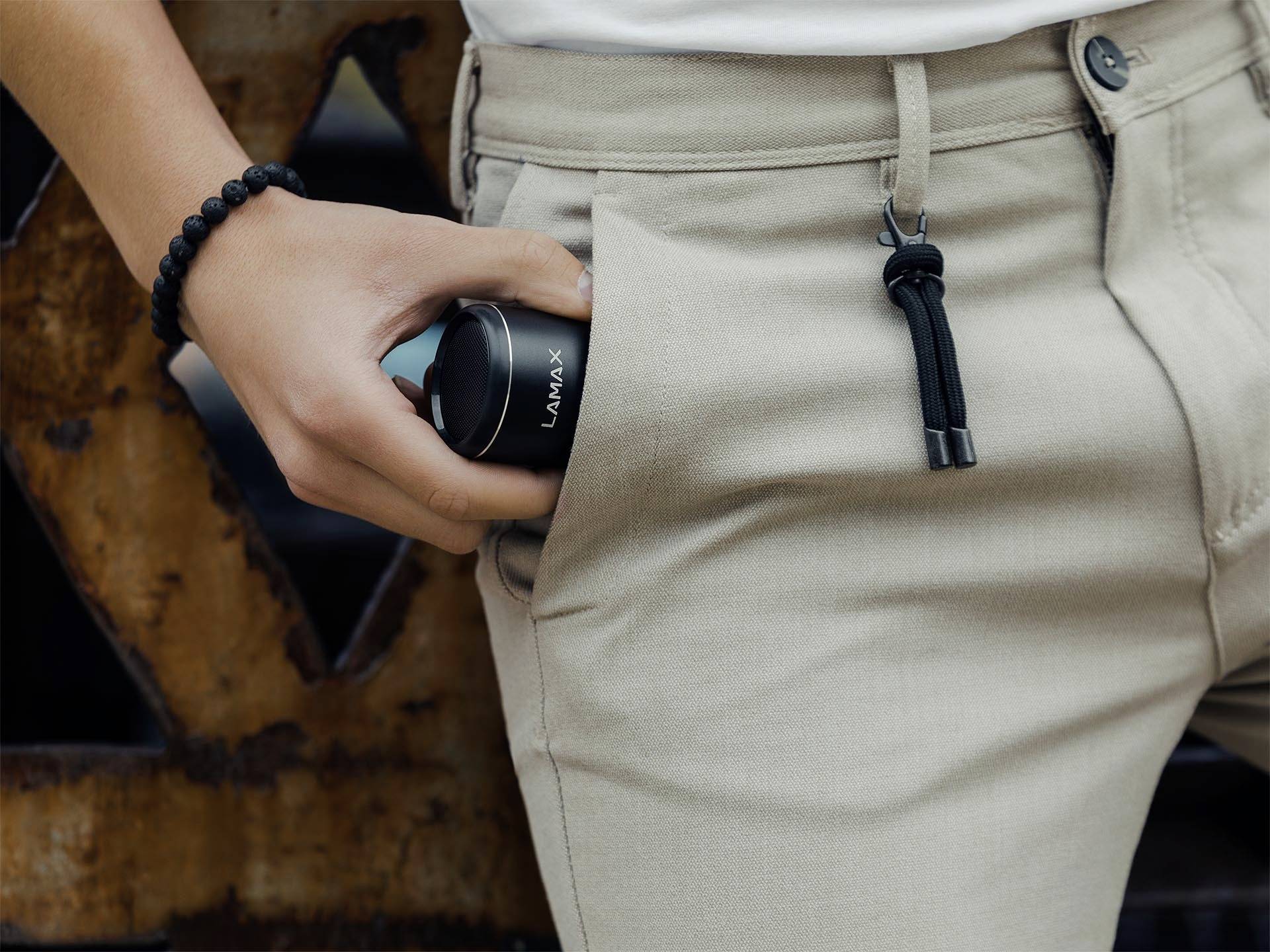 Egy ember egy kis, kerek hangszórót tesz a nadrágzsebébe. A hangszóró fekete és a "LAMAX" feliratot viseli.