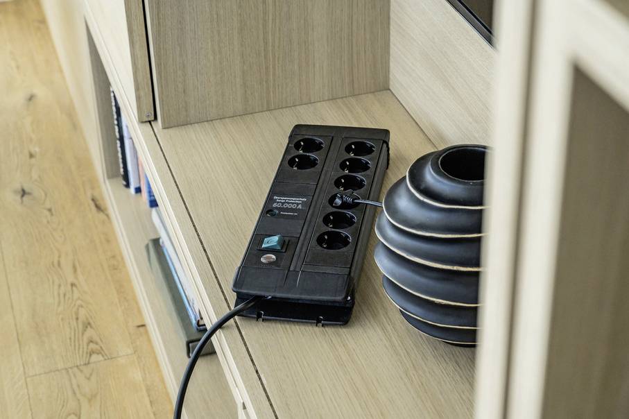 Egy fekete elosztó egy csatlakoztatott kábellel egy fa polcon fekszik egy fekete váza mellett.