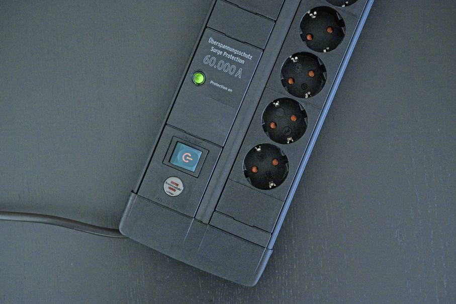 Egy fekete elosztó túlfeszültség-védelemmel és hat csatlakozóaljzattal az asztalon. Egy zöld jelzőfény mutatja az üzemkészséget.