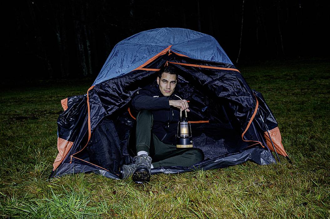 Egy férfi egy nyitott sátorban ül egy réten, sötétben, egy lámpással a kezében.