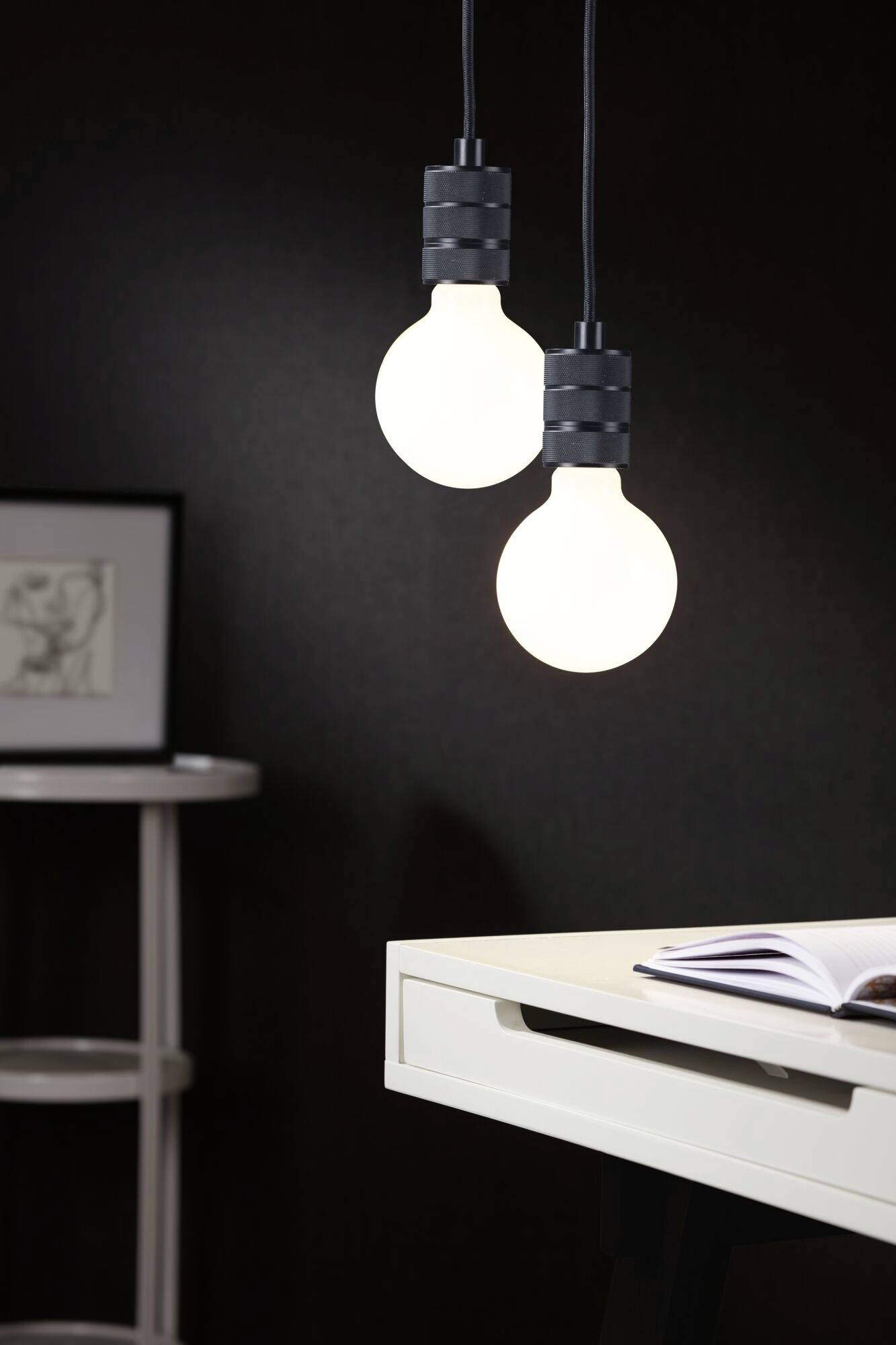 Két lógó villanykörte világít egy íróasztal fölött, amelyen egy nyitott könyv fekszik. A háttérben egy keretezett kép látható.