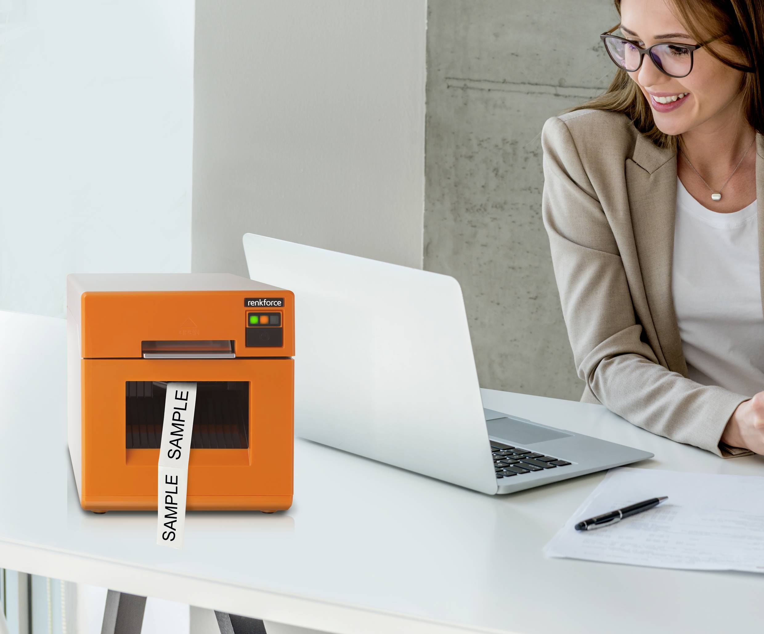 Egy nő ül egy íróasztal mellett, előtte laptop, jegyzetfüzet és toll. Mellette egy narancssárga nyomtató áll, amelyen 'SAMPLE' felirat látható.