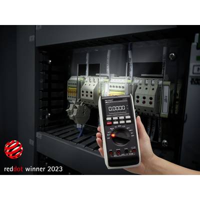 Egy kéz digitális multimétert tart egy elektromos alkatrészeket tartalmazó elosztószekrény előtt, a '2023-as Red Dot díj' jelzés a sarokban.