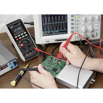 Egy személy egy zöld nyomtatott áramkört (PCB-t) vizsgál multiméterrel, miközben egy oszcilloszkópon szinuszos hullámforma látható.