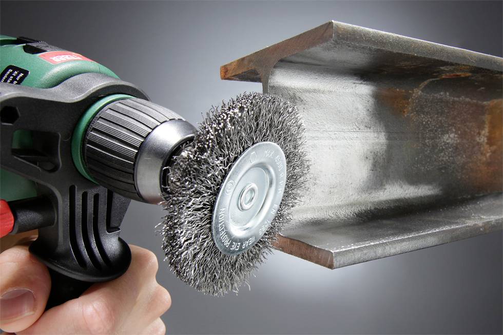 Egy kéz egy drótkeféhez tartozó sarokcsiszolóval tisztít egy acélgerendát, rozsdát és törmeléket távolítva el róla.