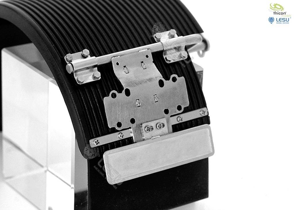 Fémes mechanizmus összetett részletekkel, egy hajlított, strukturált szalagon felszerelve. A háttérben egy kocka látható.