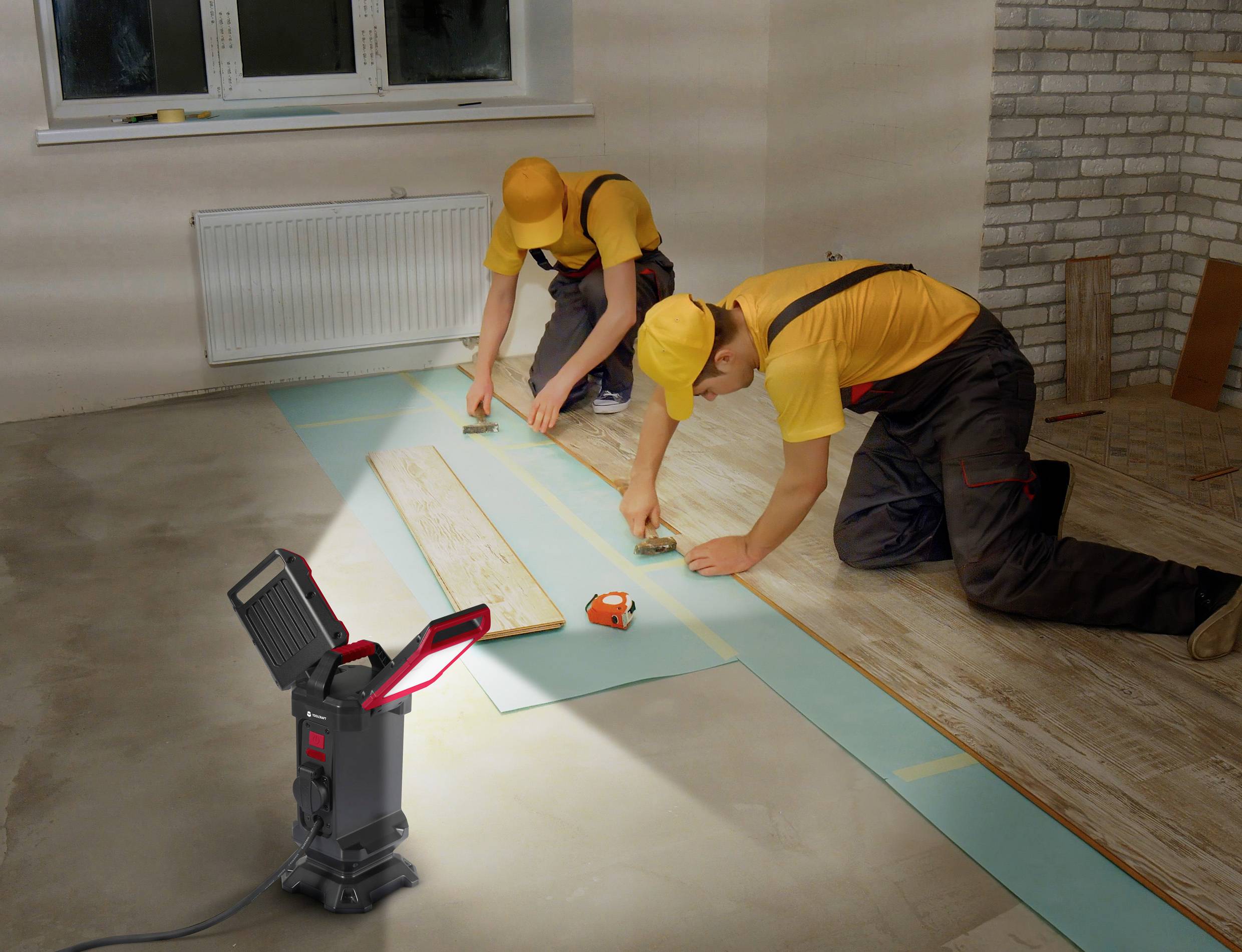 Két munkás laminált padlót fektet le egy helyiségben. Az egyik mér egy mérőszalaggal, míg a másik igazítja a laminát elemeket. Egy lámpa világítja meg a területet.