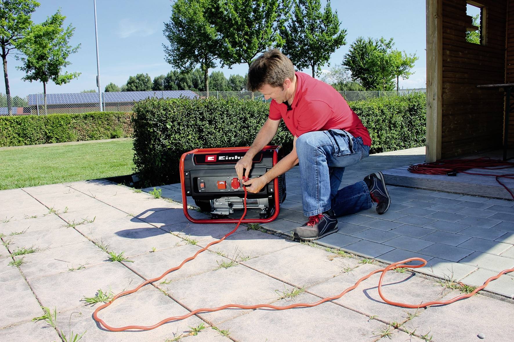 Egy férfi piros pólóban térdel egy teraszon, és egy hordozható generátorhoz csatlakoztat egy elektromos kábelt. A háttérben fák láthatók.