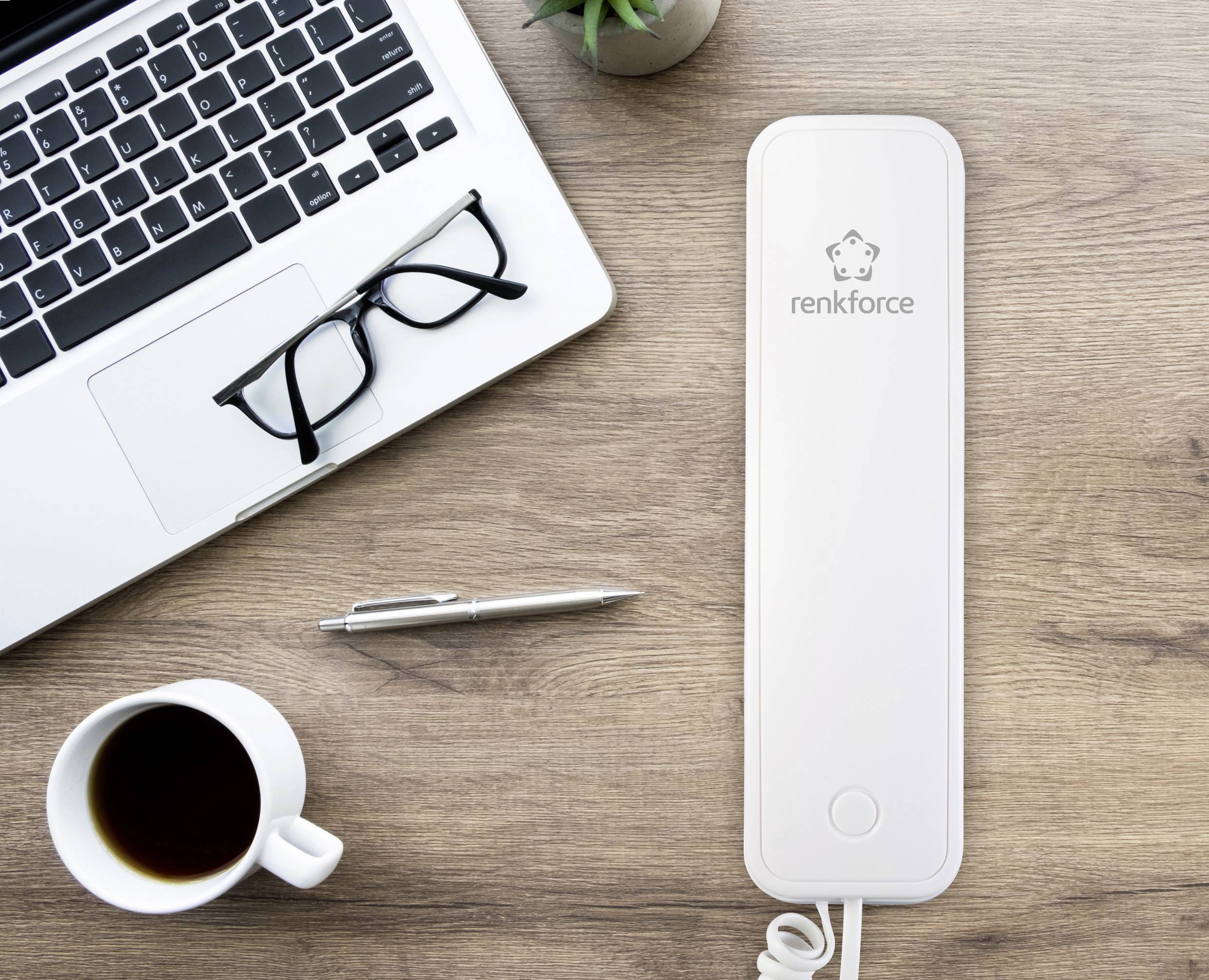 Egy íróasztal laptoppal, szemüveggel, kávésbögréval, tollal és egy fehér, téglalap alakú eszközzel, amelyen a 'Renkforce' felirat szerepel.