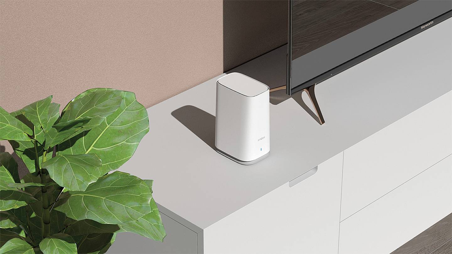 Egy fehér WiFi-router áll egy fehér szekrényen egy televízió mellett. Baloldalon egy zöld növény látható.