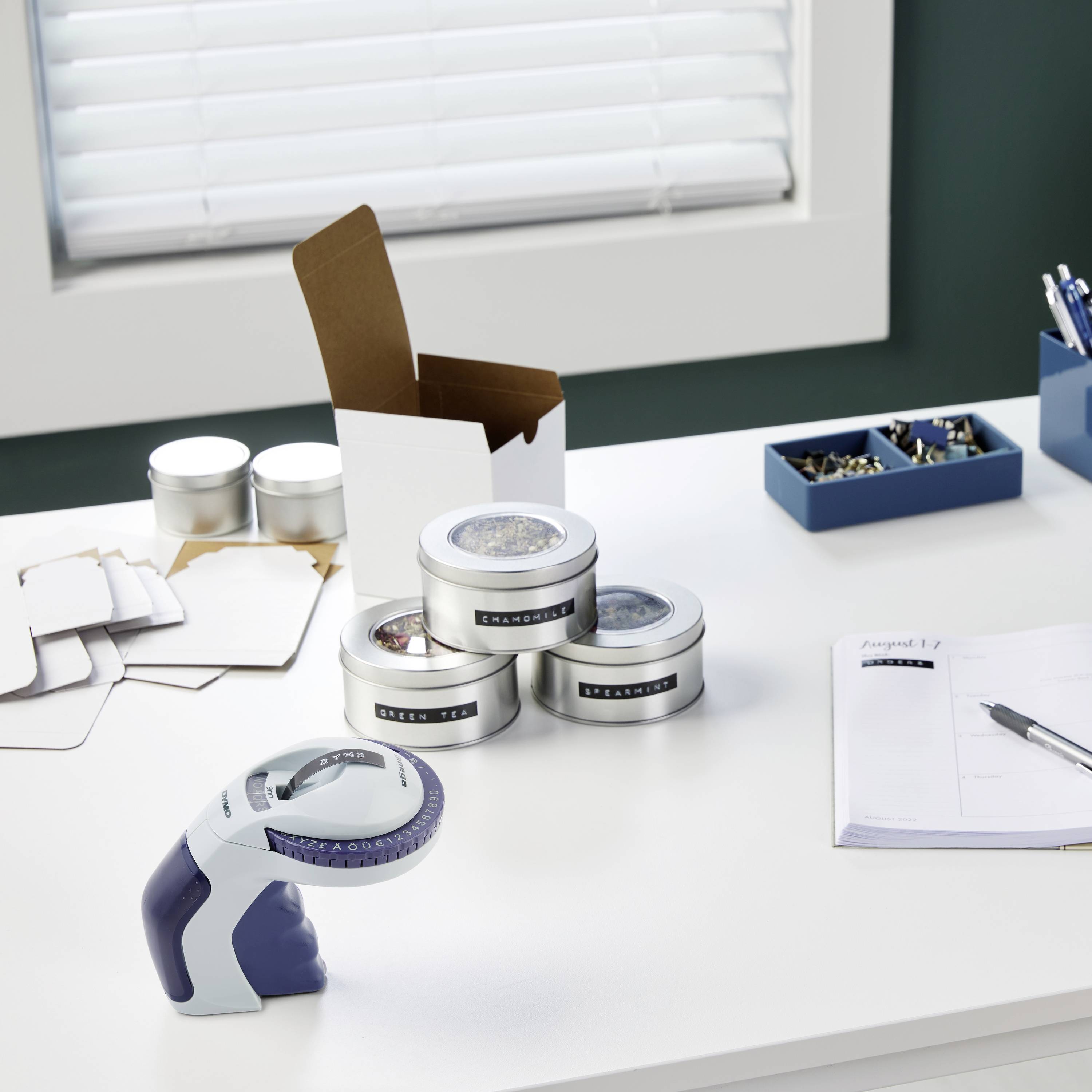 Munkasztal egy címkézővel, három dobozzal, kártyákkal és egy jegyzetfüzettel egy ablak alatt. Egy kisebb kartondoboz nyitva van.