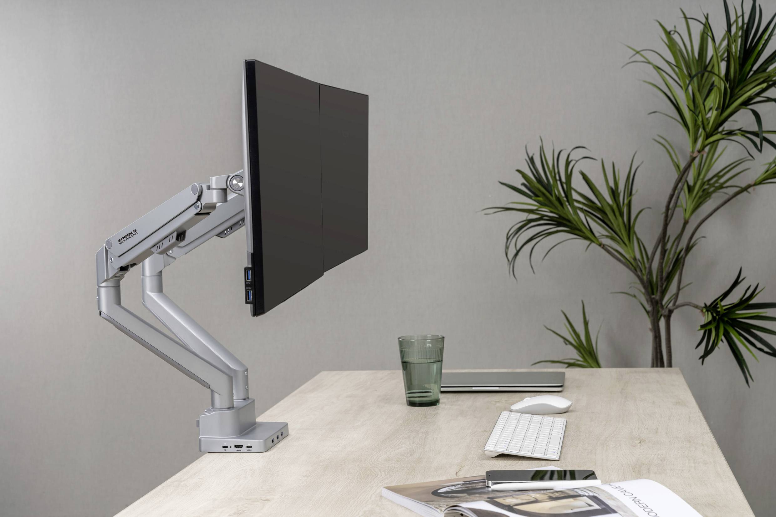 Egy modern munkahely magasságállítható monitorkarral, monitorral, billentyűzettel, egérrel és növényekkel a háttérben egy fa asztalon.