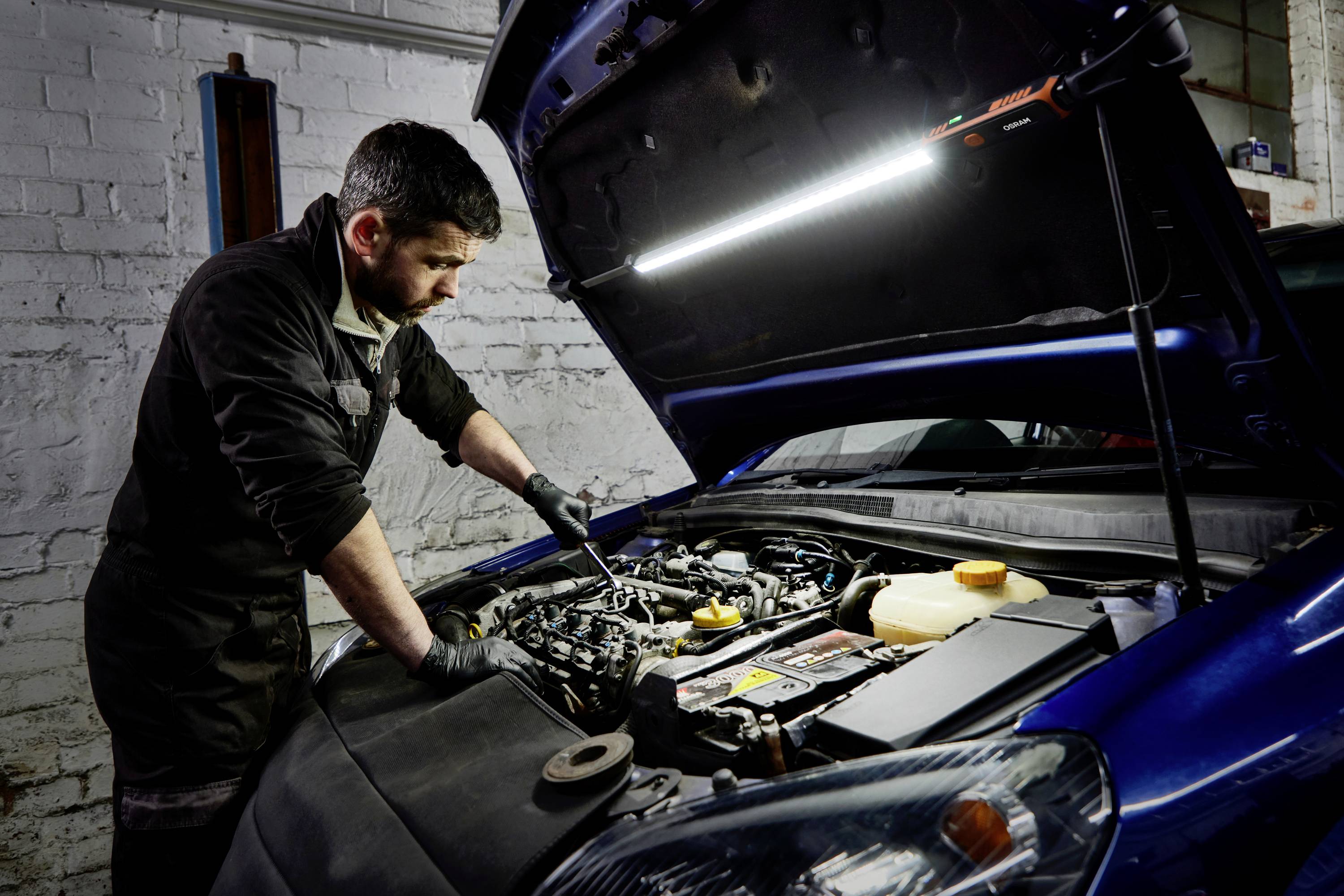 Egy szerelő motor javítását végzi egy nyitott motorháztető alatt egy műhelyben, egy LED-lámpa megvilágításában.