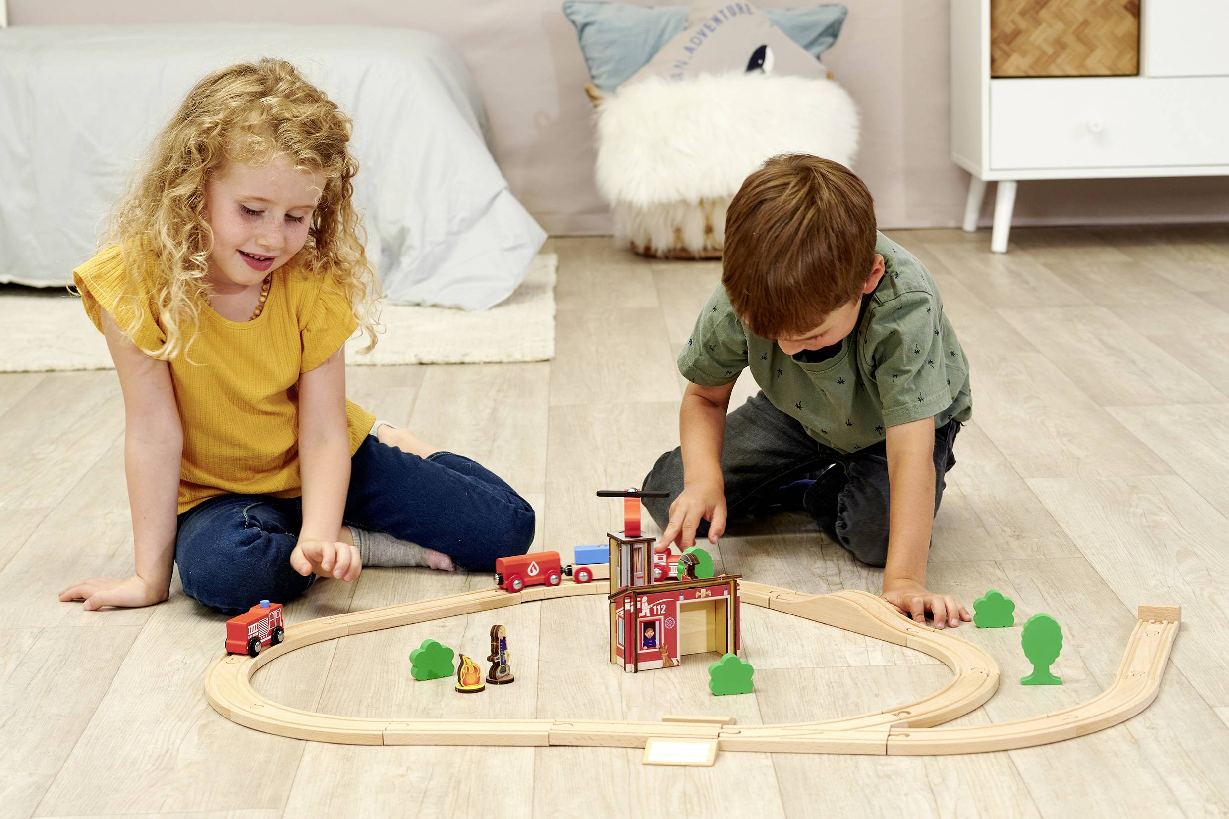 Két gyerek játszik a padlón egy fa vonattal. Egy fiú és egy lány tolja a vonatokat egy kis fákkal tarkított pálya mentén.