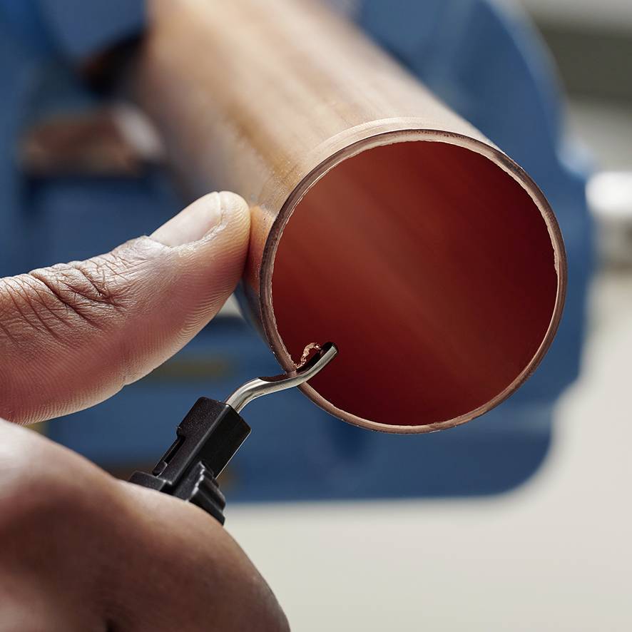 Egy kéz egy rézcsövet tart, amelyet egy sorjázóval munkálnak meg, hogy eltávolítsák a éles széleket.