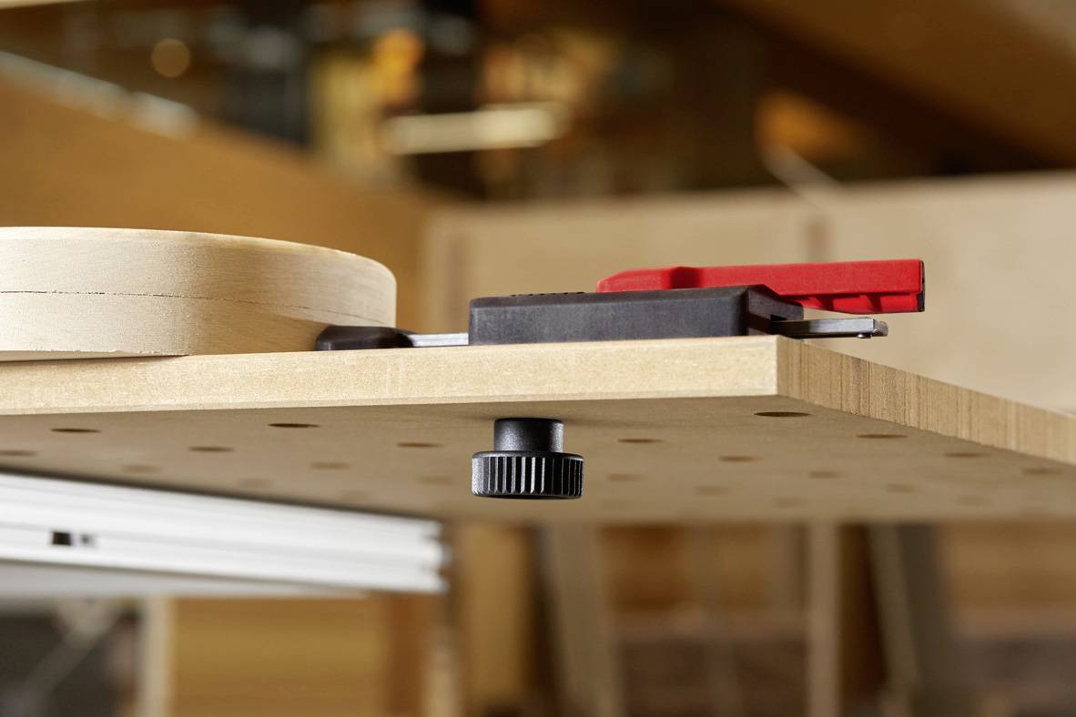 Egy fa munkaasztal állítható szorítórendszerrel és lyukas lemezzel. A felületen körlapok és egy piros szorító található.