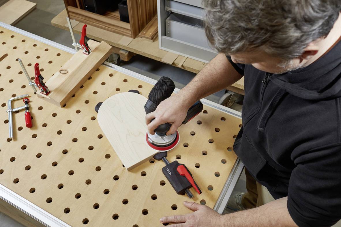 Egy férfi egy szívformájú fadarabot csiszol egy lyukasztott táblán egy csiszológéppel, szerszámok körülvételében.