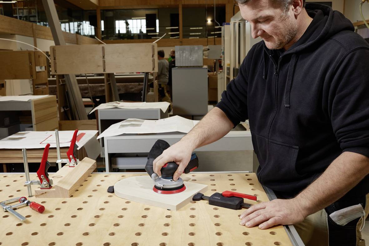 Egy férfi egy munkaasztalon egy fadarabot csiszol egy műhelyben. Körülötte szerszámok és más fafaragványok vannak.