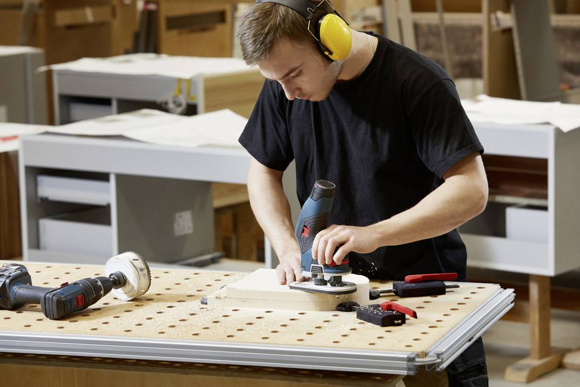 Egy férfi egy fűrésszel dolgozik egy falemezen egy műhelyben. Védőfülest visel, és szerszámok veszik körül.