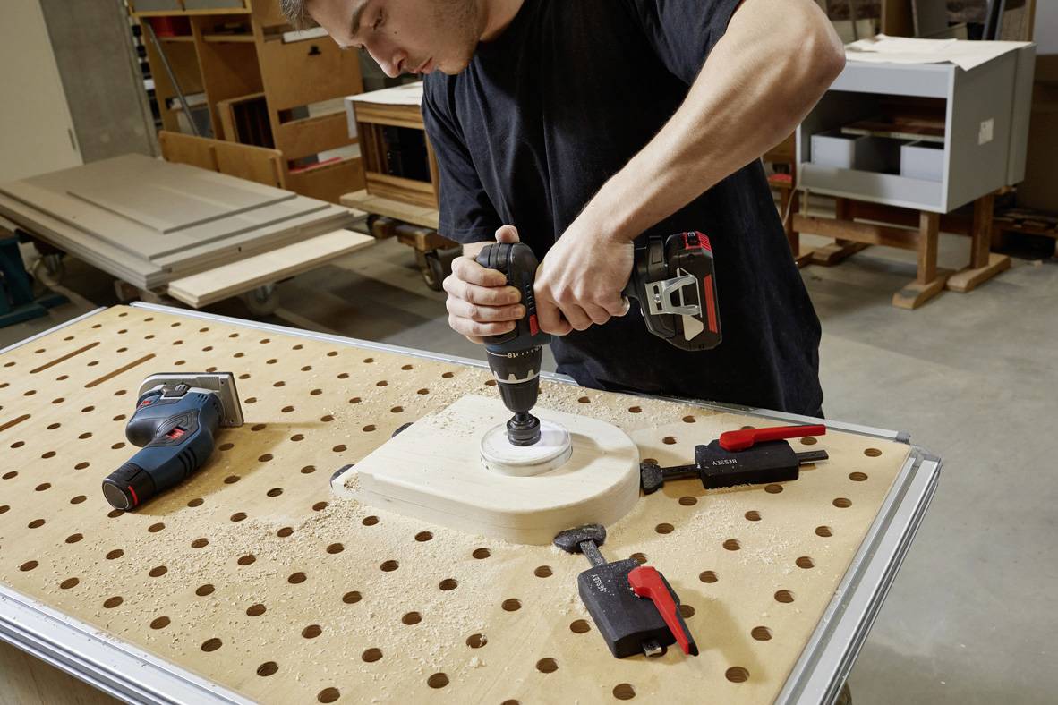 Egy férfi egy műhelyben lyukat fúr egy fadarabba, amely egy munkaasztalon rögzítve van szorítófogókkal.