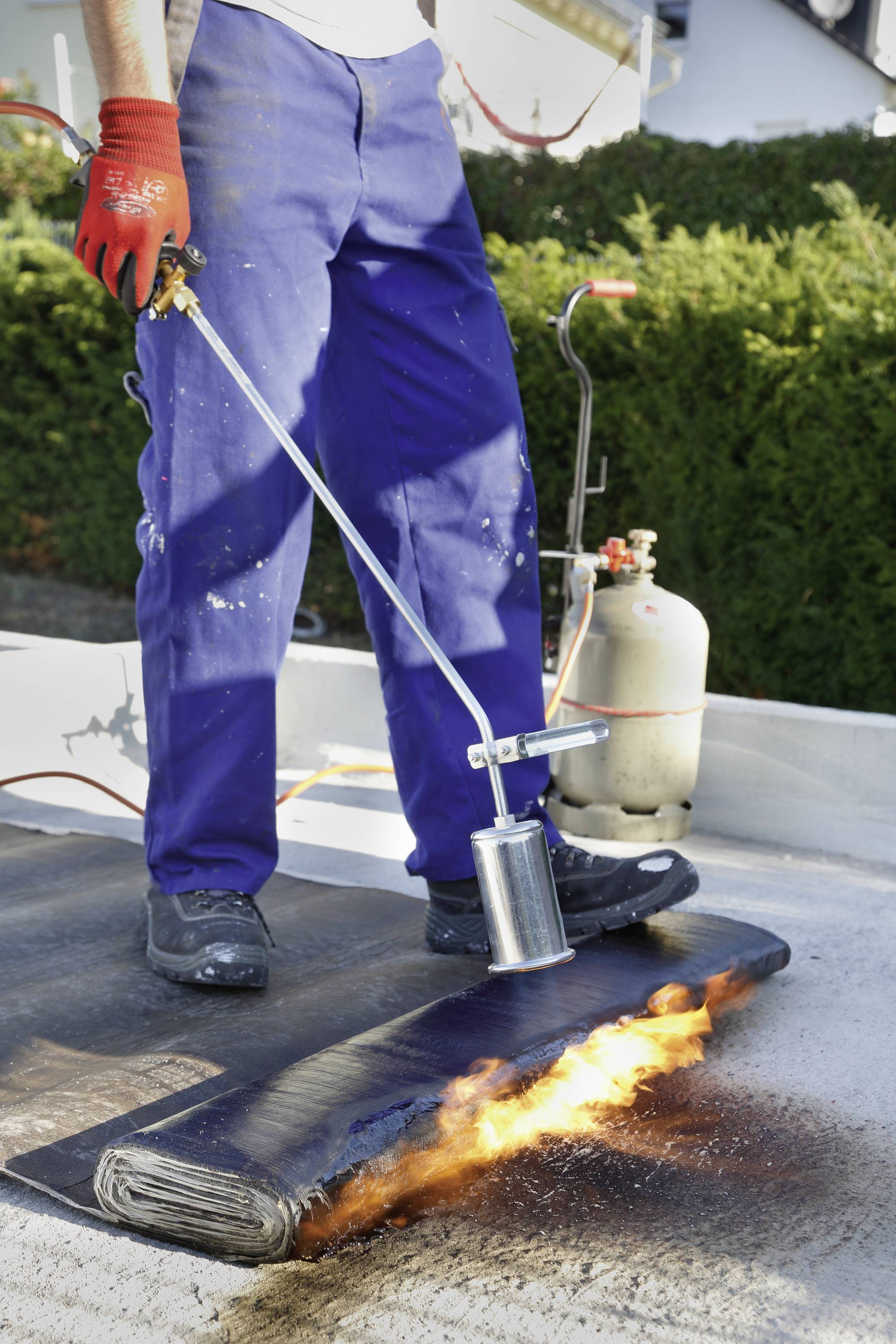Egy személy egy tetőn áll, gázégővel hevíti a tetőszigetelő szalagot. Biztonsági felszerelés és gázpalack látható.