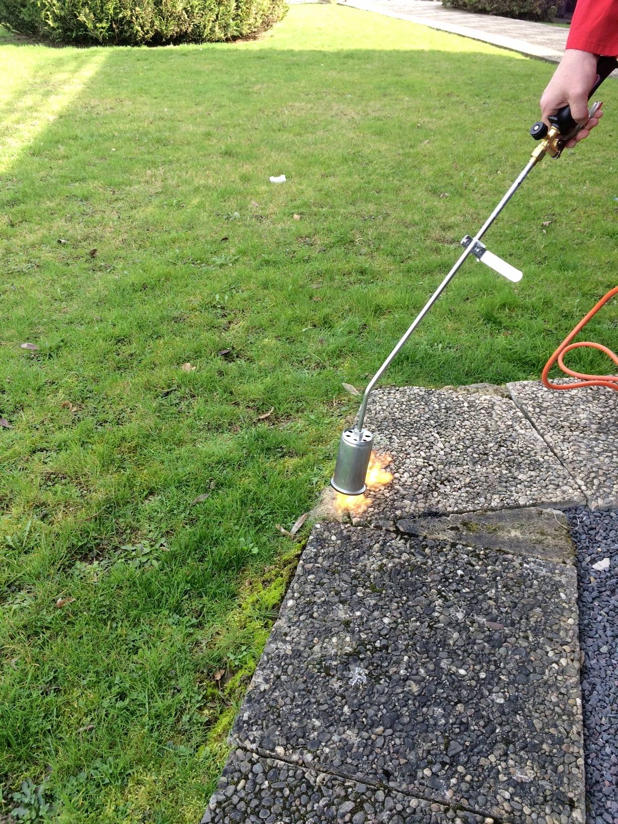 Egy személy gázlángfejlesztőt használ, hogy eltávolítsa a gyomot a járólapok között egy füves területen.
