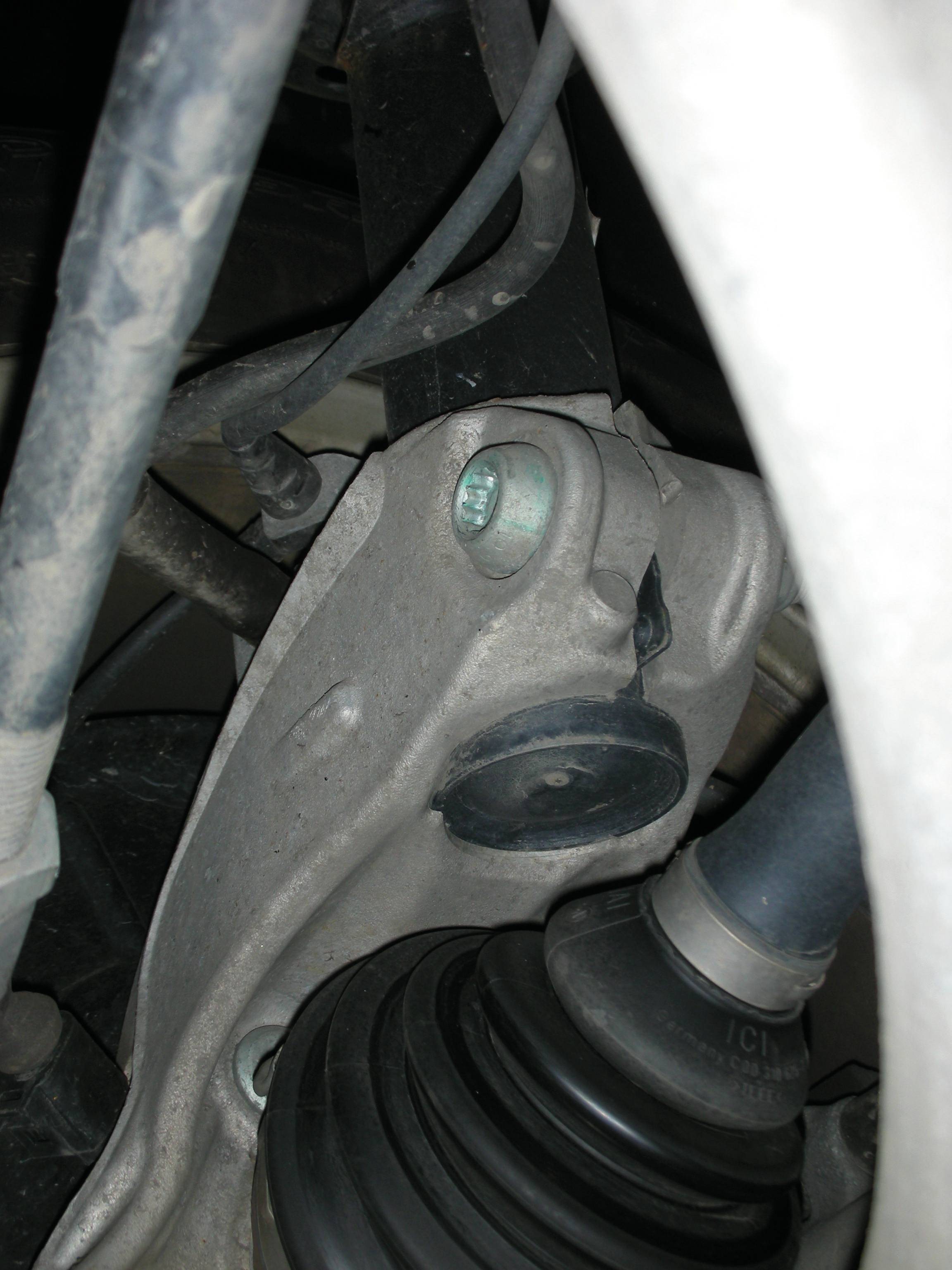 Egy autóalkatrész közeli felvétele, valószínűleg a futómű vagy a kerékfelfüggesztés egy része, amelyen egy lengéscsillapító, egy tartókonzol és egy tengely látható.