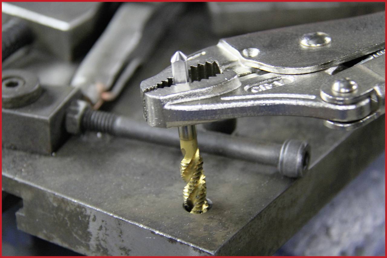 Egy fogó tart egy menetfúrót, amelyet egy fémdarabba csavarnak, hogy meneteket alakítsanak ki. A háttérben szerszámok láthatók.