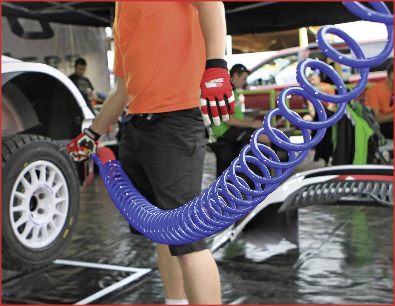 Egy személy egy nagy, kék spirál tömlőt tart a kezében, feltehetően egy műhelyben vagy egy járműverseny helyszínén.