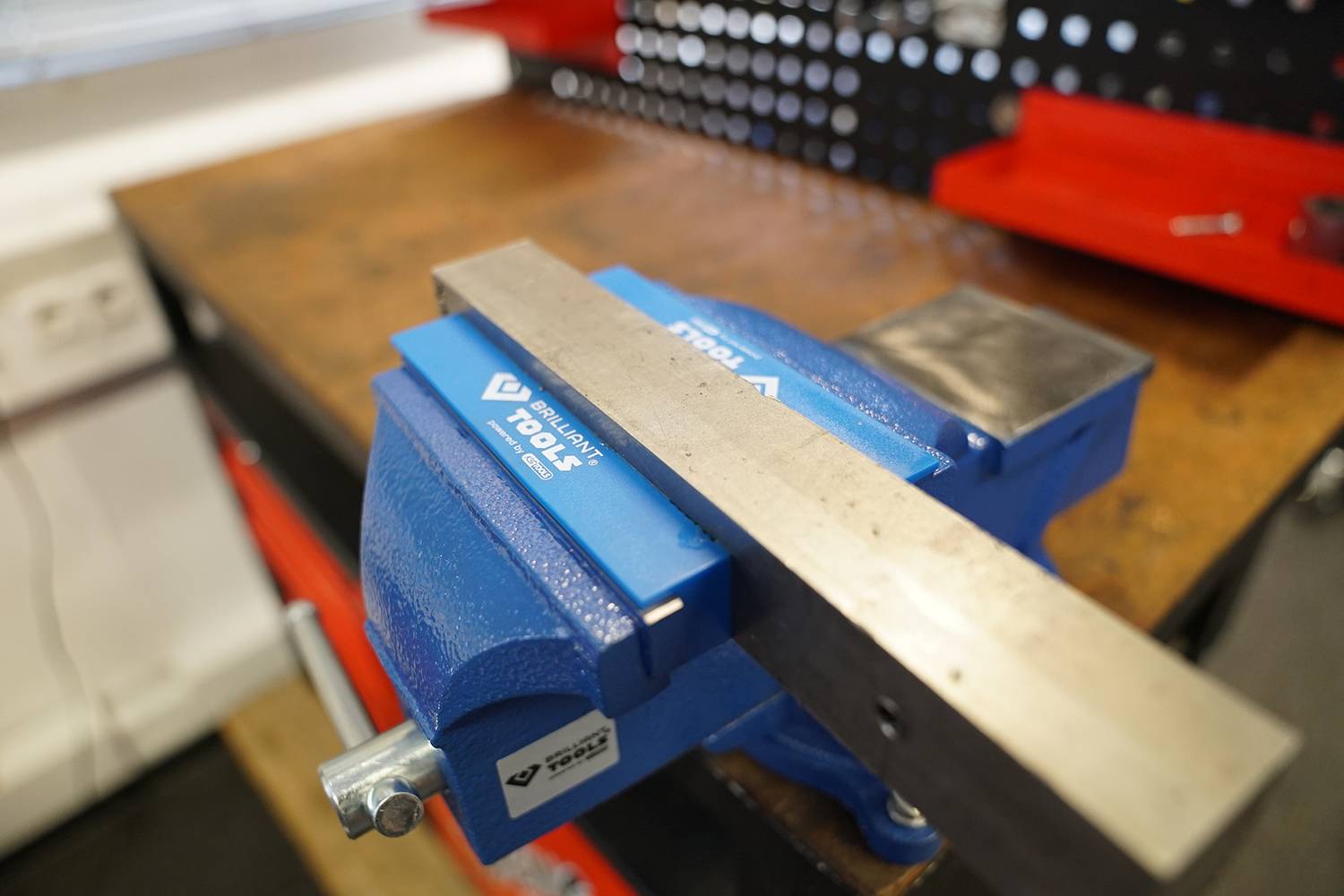 Egy satuba szorított fémdarab egy munkapadon. A háttérben piros szerszámos szekrények láthatók.