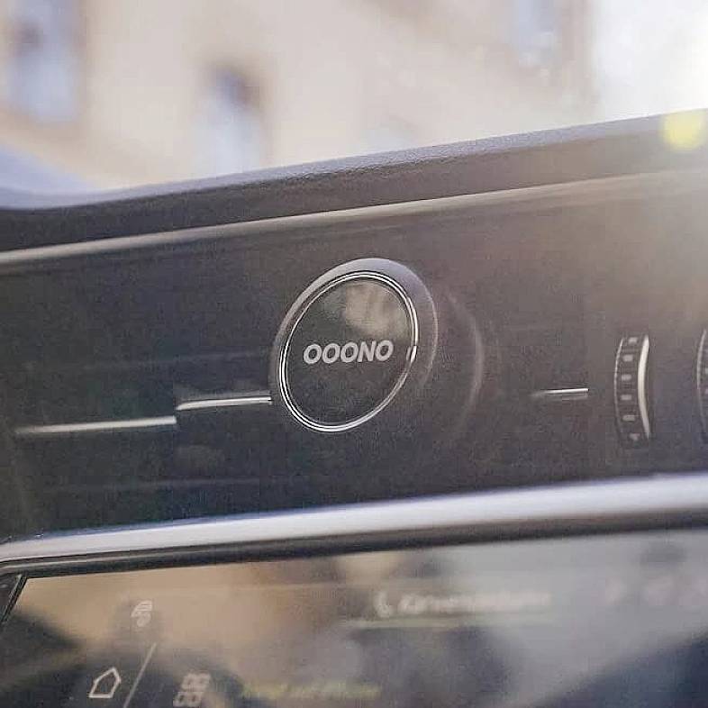 Egy OOONO feliratú kerek gombbal rendelkező automatikus rendszer az autóban. A háttérben az autó belseje és elmosódott külső látható.