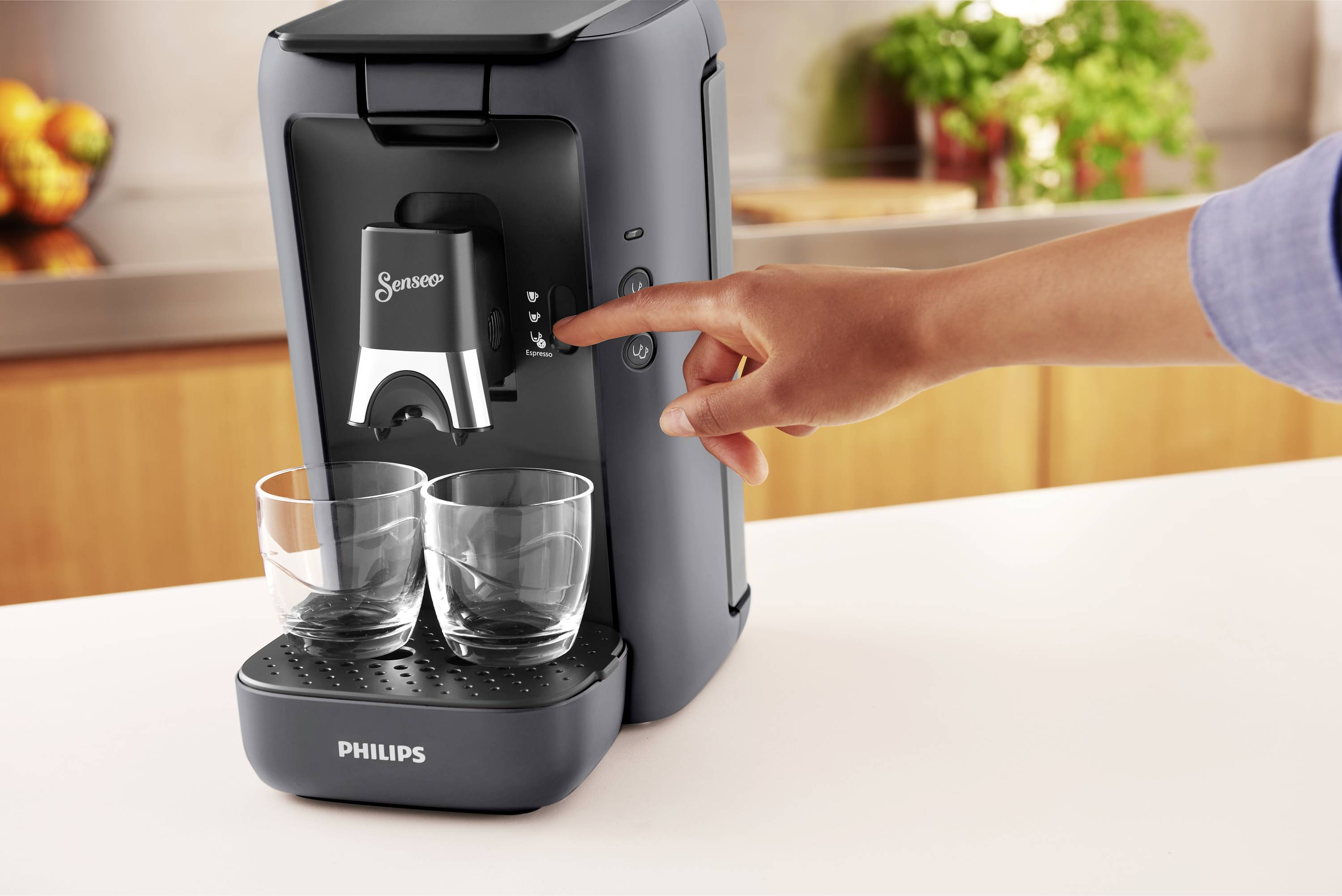 Egy személy egy kávégépet kezel, amelynek két üres pohár van alatta. A háttérben egy konyhapult gyümölcsökkel.