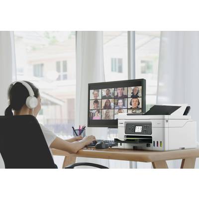 Egy személy ül az íróasztalánál fejhallgatóval, és részt vesz egy videókonferencián a számítógép képernyőjén. Jobbra mellette egy nyomtató áll.