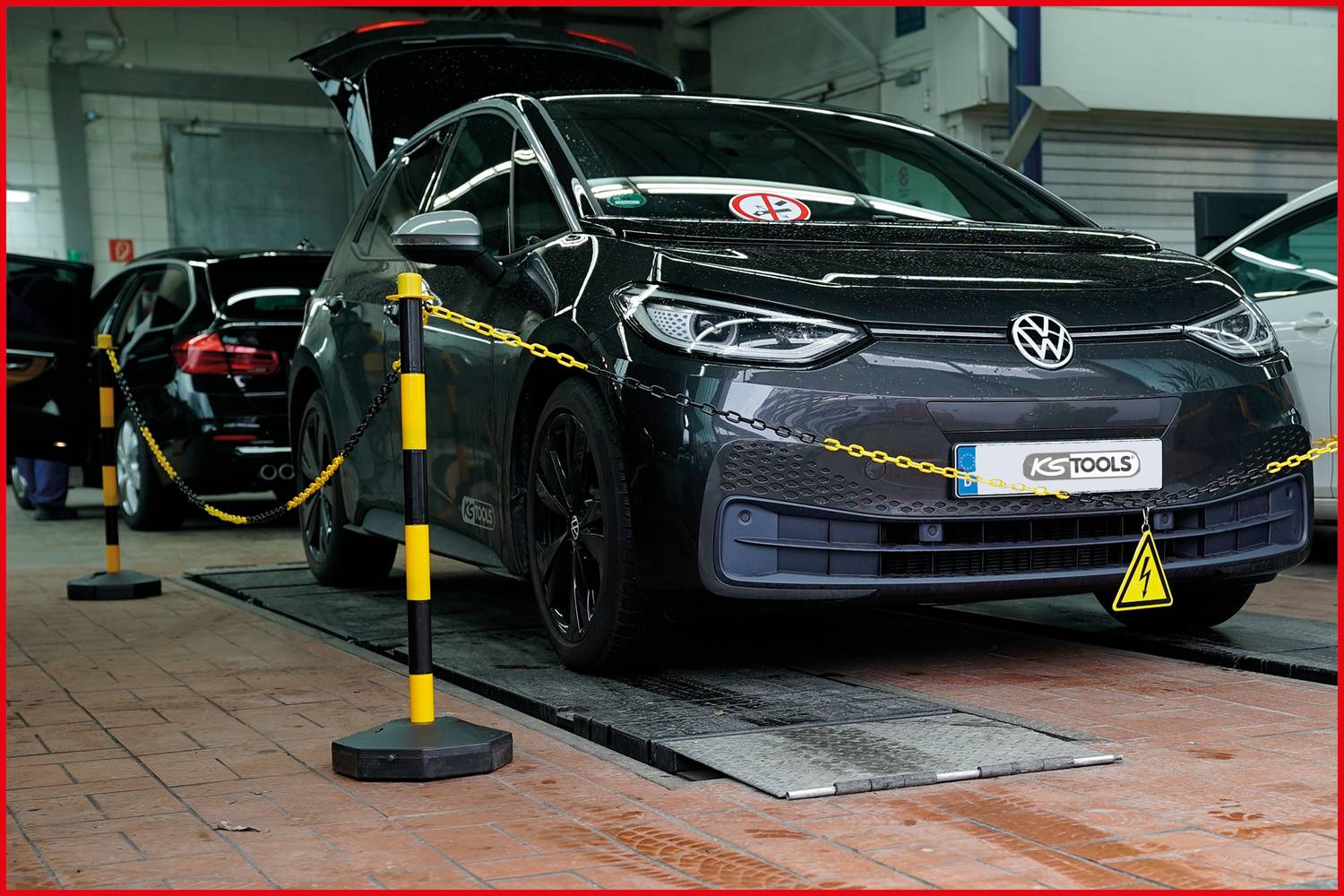 Egy fekete Volkswagen nyitott csomagtérrel egy autószerelő műhelyben áll egy emelőn. Biztonsági láncok és táblák láthatók.