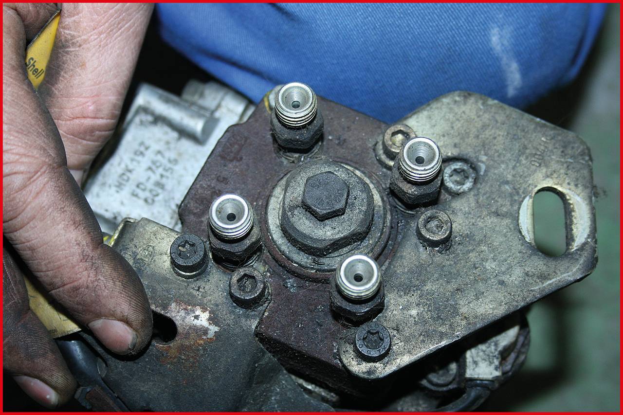 Egy kéz egy mechanikai alkatrészt tart, amelyen több csavarkapcsolat látható. A háttérben egy kék ruhadarab figyelhető meg.