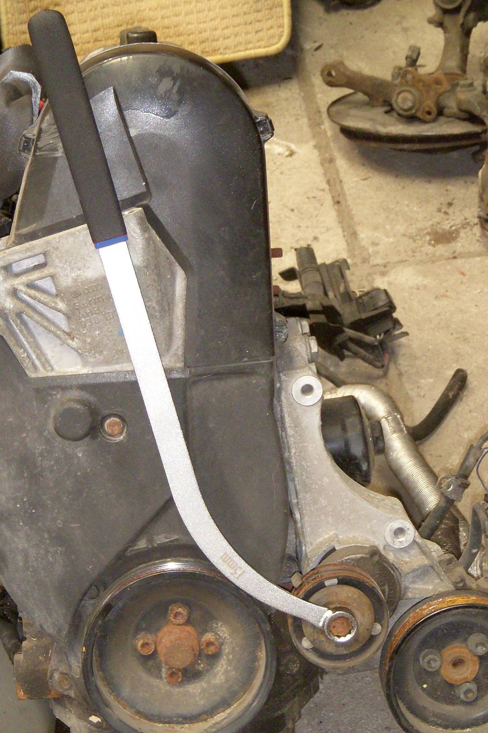 Egy hosszú nyélű szerszám van egy autómotor alkatrészére helyezve, hogy mechanikai munkálatokat végezzenek. A háttérben további autóalkatrészek fekszenek.