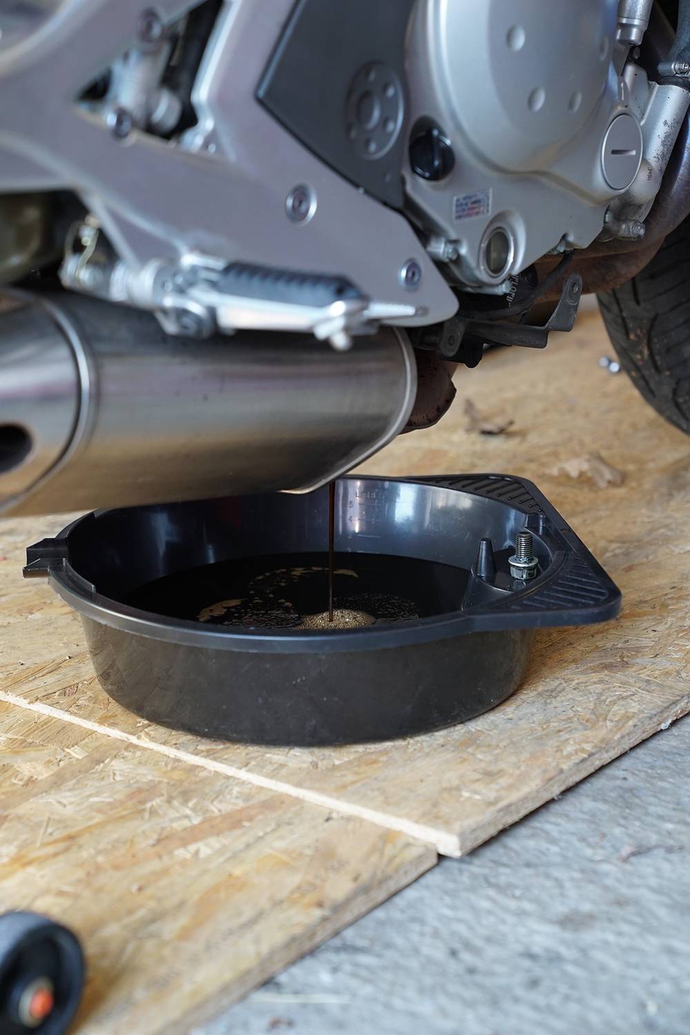 Olaj folyik egy motorkerékpárból egy gyűjtőedénybe. A jelenet egy olajcserét mutat be fa padlóburkolaton.