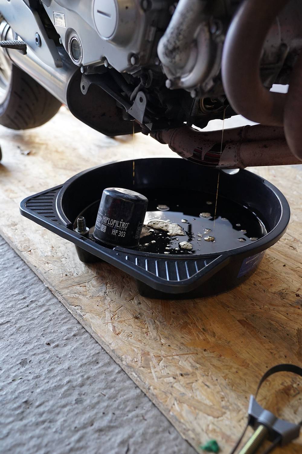 Motorolaj-csere egy motorkerékpárnál: A használt olajszűrő és a régi olaj egy fekete felfogótálcába folyik a motor alatt.